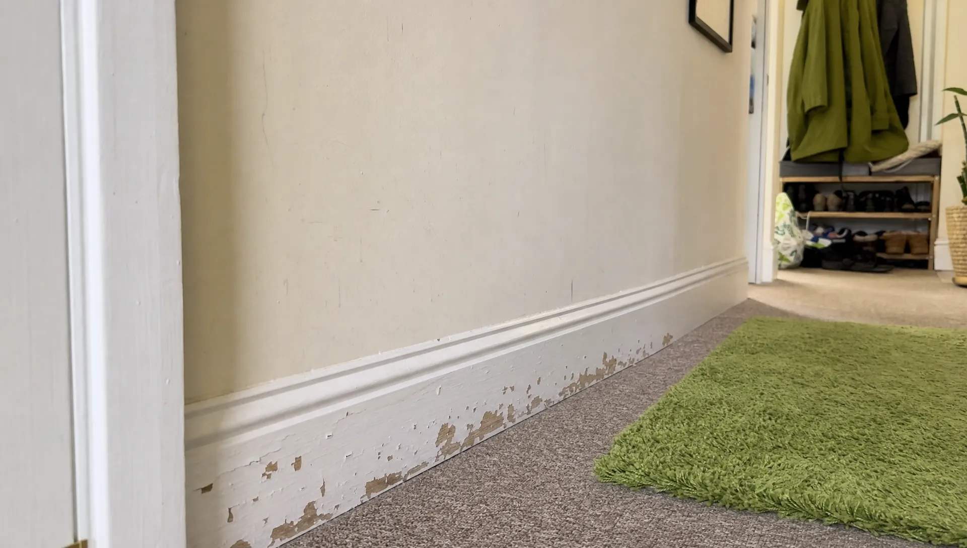 A normal-height view of a British hallway showing white skirting boards with visible flaking and peeling paint near the floor, with a slightly scuffed wall, carpet edge, and everyday household clutter in the background.